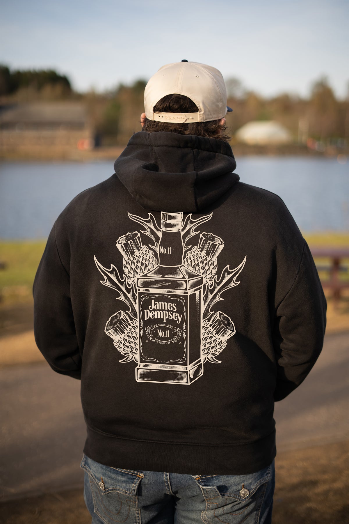 A person viewed from behind wearing a black hoodie with a large white James Dempsey bottle graphic on the back, a white baseball cap, and blue jeans, standing outdoors near a lake.